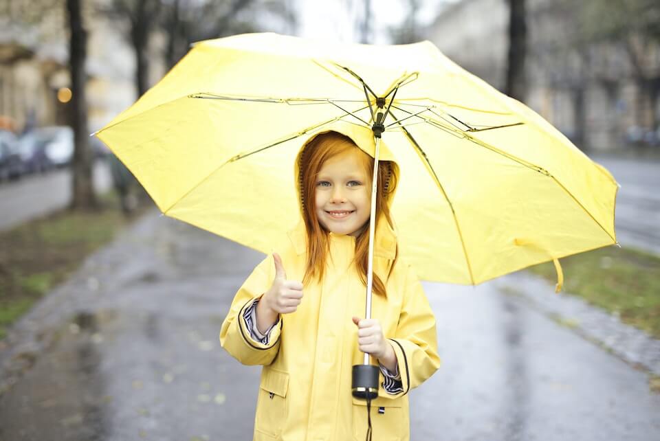 Little girl wearing a yellow raincoat and holding a yellow umbrella while giving a thumbs up