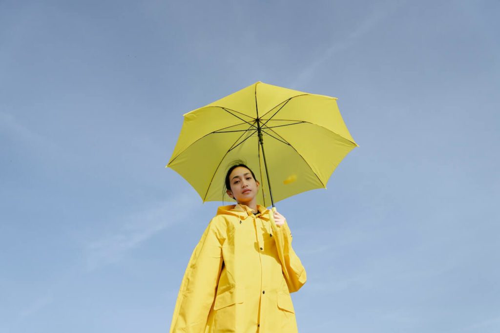 Woman wearing yellow raincoat and holding yellow umbrella while standing under a clear blue sky